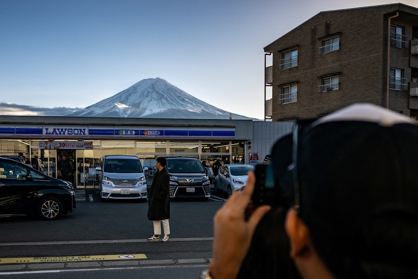 A man poses in front of a service station with Mt Fuji in the background as someone takes a photo