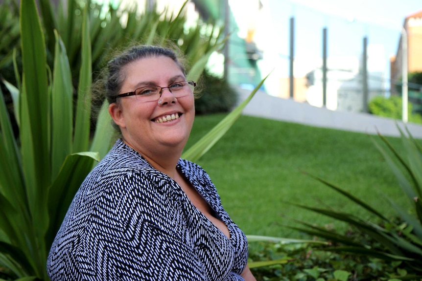 Debbie, a woman with glasses and a nose ring, sits smiling in a park, surrounded by greenery.