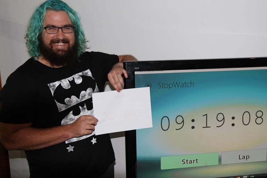 Man with blue-green hair stands in front of a timer and holds a piece of paper.