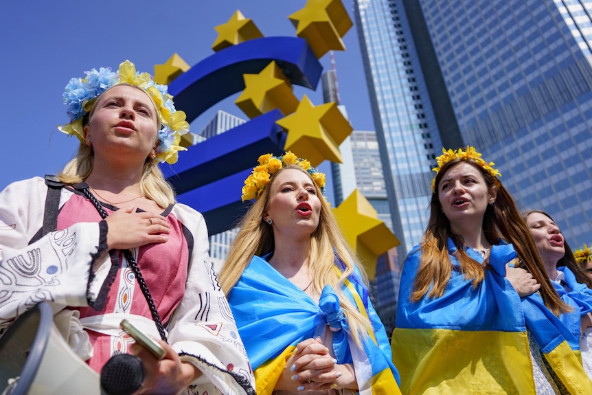 Trois femmes portant des drapeaux ukrainiens bleus et jaunes avec des fleurs jaunes dans les cheveux lors d'une manifestation. 