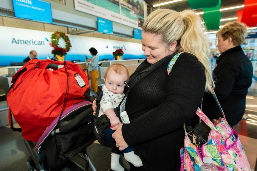 A mum holds a baby in a chest carrier in line for check-in at an American airline