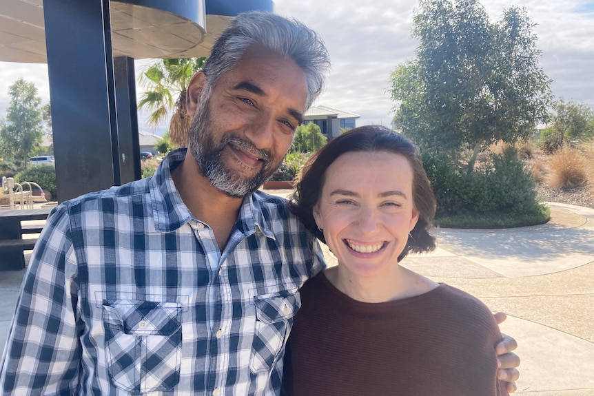 A man in a checked shirt with his arm around a woman in a brown jumper both smiling at the camera