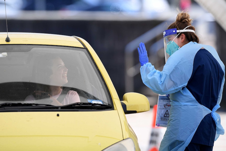A woman in a yellow car and a COVID testing woman smile at each other