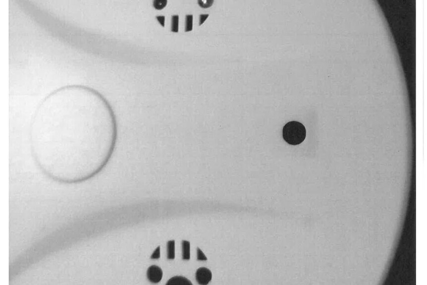 A close up shot shows a tiny lens on a white smoke detector