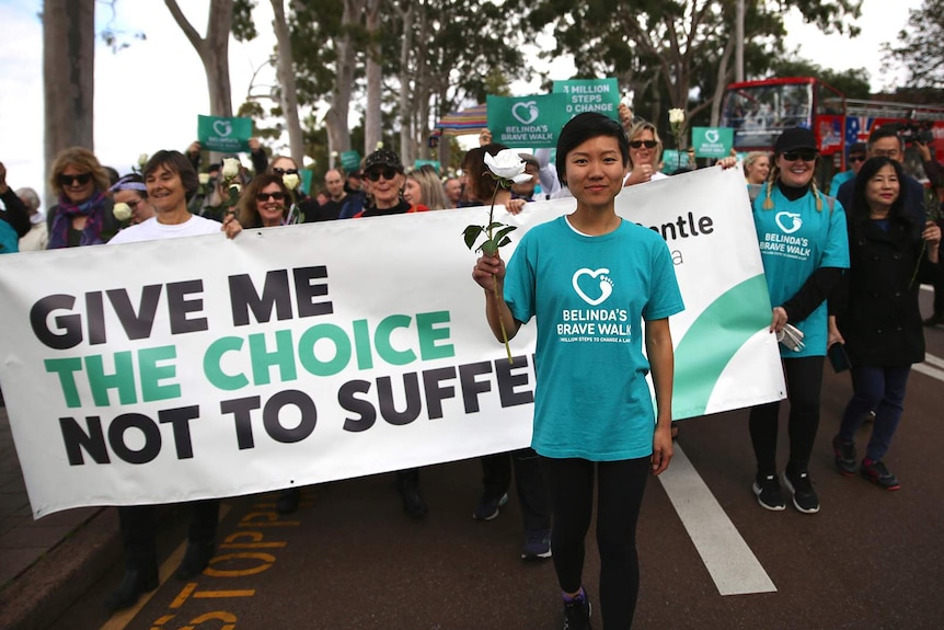 Belinda Teh poses for a photo at Kings Park in Perth holding a white rose and a photo while smiling.