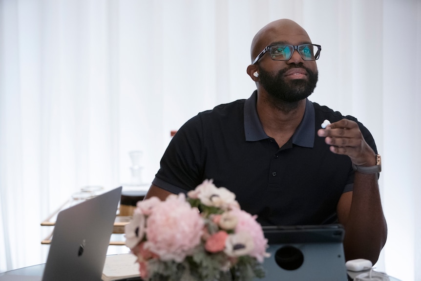 A Black person in their 30s with glasses and a beard and a dark polo shirt sits a desk with flowers and a laptop