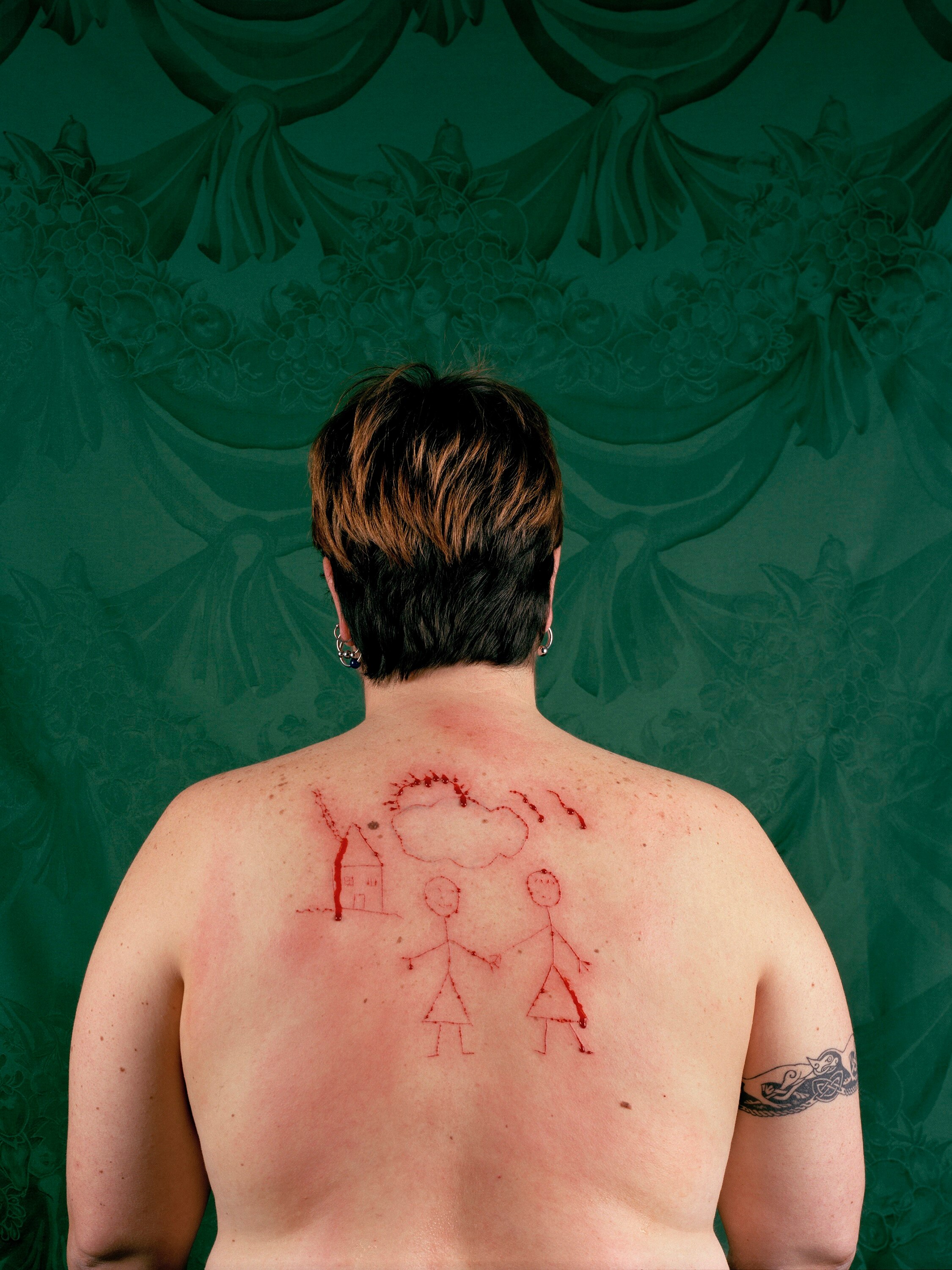 A woman has short hair, a tattoo on her right shoulder and a drawing of a lesbian family cut into the bare skin of her back