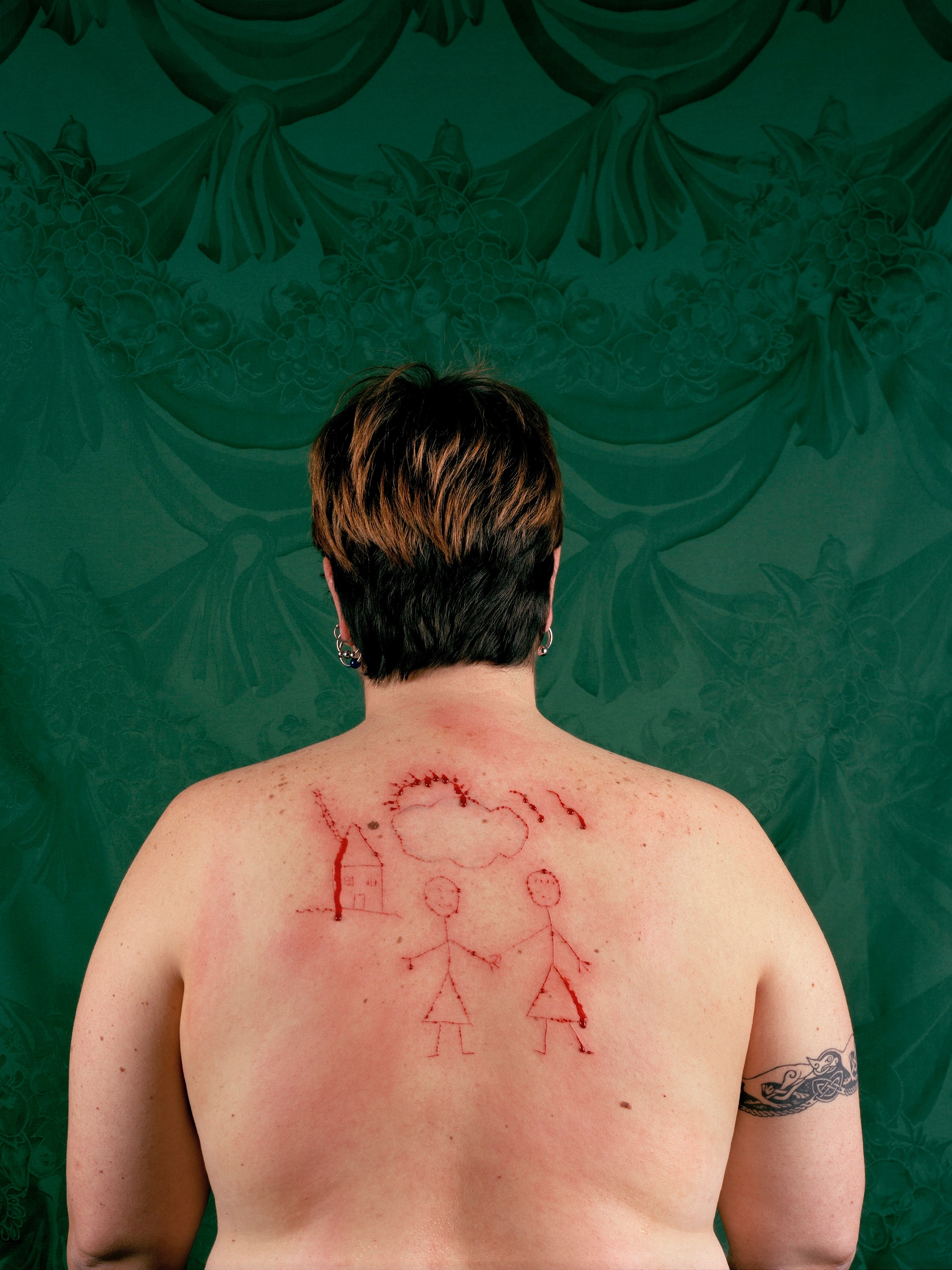 A woman has short hair, a tattoo on her right shoulder and a drawing of a lesbian family cut into the bare skin of her back