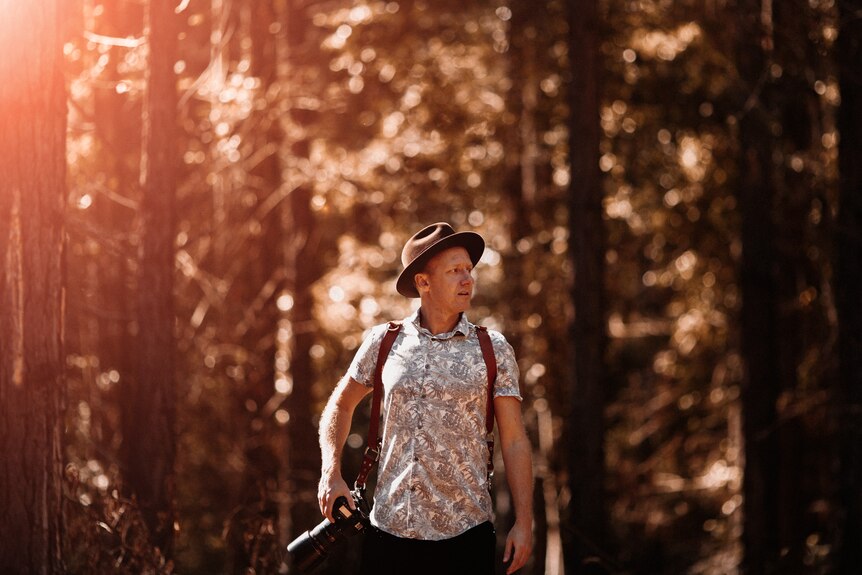 middle-aged man in a casual shirt carrying gear on his back, walking through a forest with a camera over his shoulder