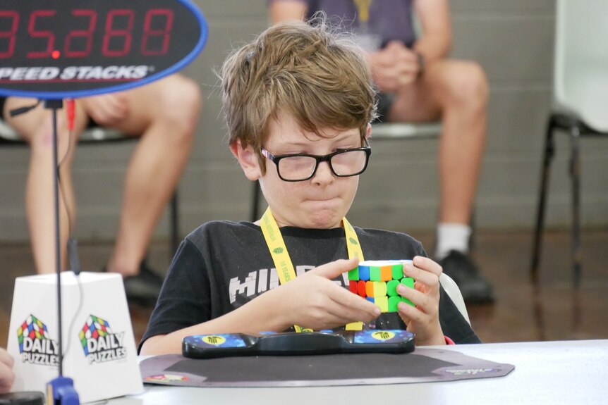 A boy in a black shirt sits at a table holding a Rubik's cube