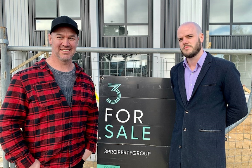 Two men stand in front of a for sale sign at a home under construction.