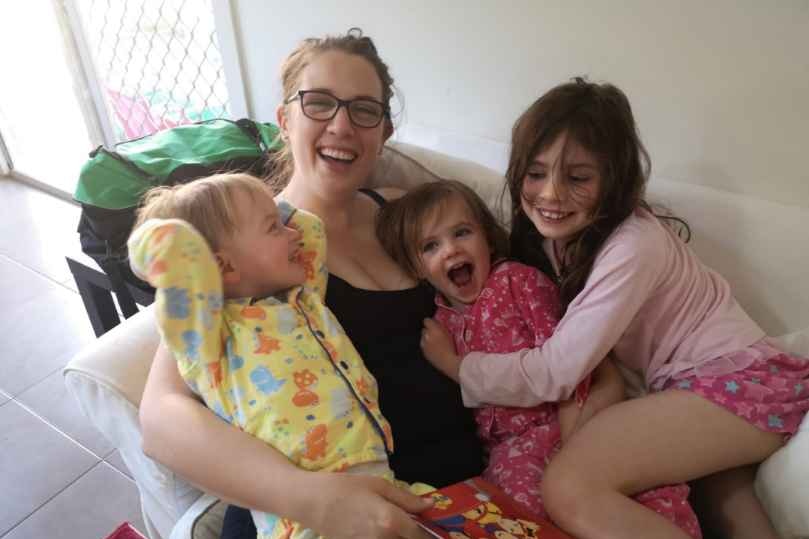 A smiling woman sits on a couch with her three children who are all laughing.