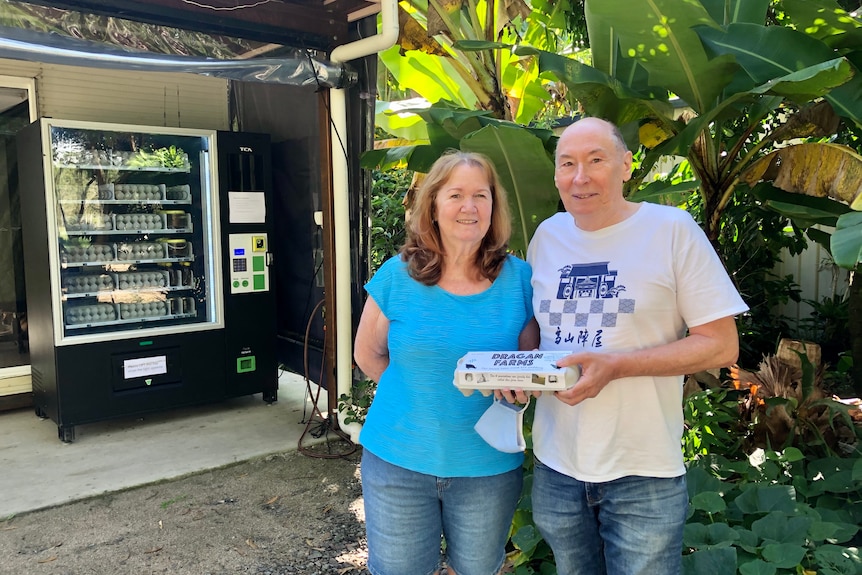 A couple wearing stand in front of the vending machine holding a carton of eggs.