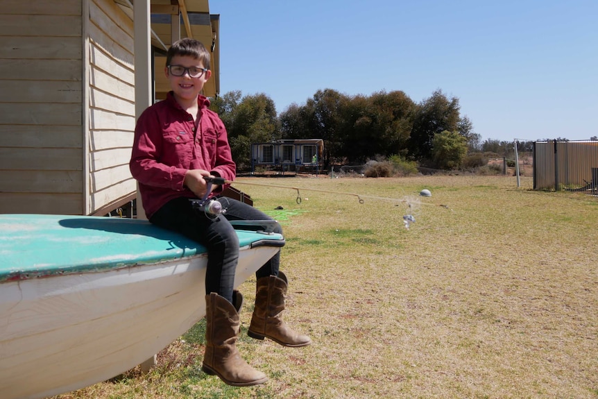 A child in a red shirt sits on the bow of a boat propped up in a front yard holding a fishing rod.