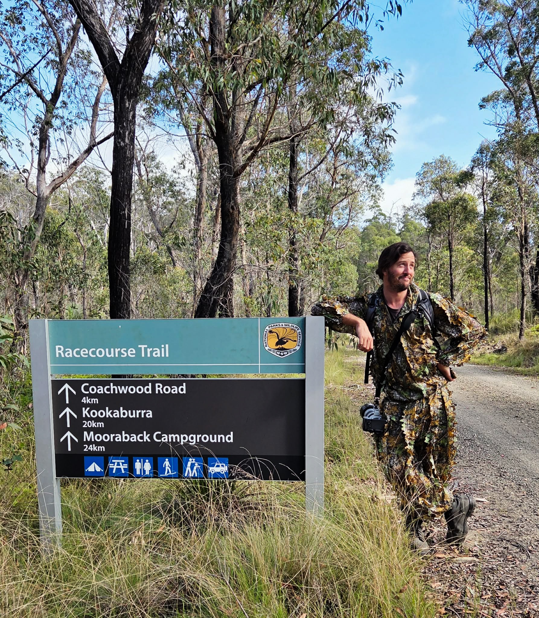 James está parado junto a un cartel en un parque nacional, vestido con ropa de camuflaje, sonriendo y mirando los árboles.