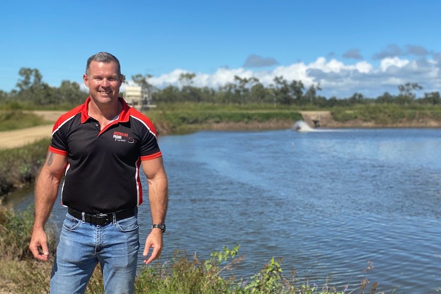 Prawn farmer Matt West stands in front of a pond on his prawn farm, trees and a road are visible in the background.