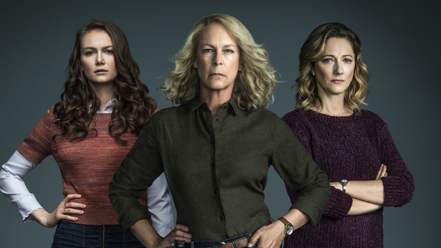 Three women stand with hands on hips or arms folded, and hair out, with solid background behind.