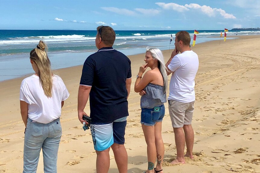 Four adults stand looking at the surf in a reflective way.