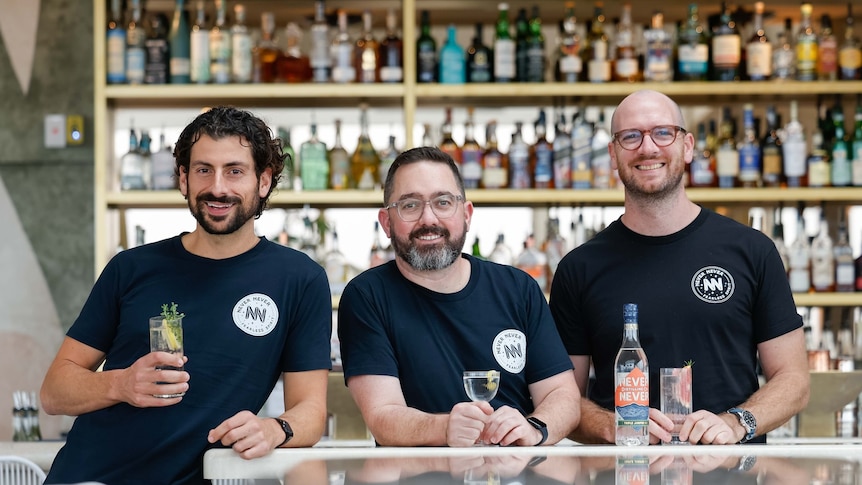 three men in black t-shirts smiling and holding alcoholic drinks behind a bar