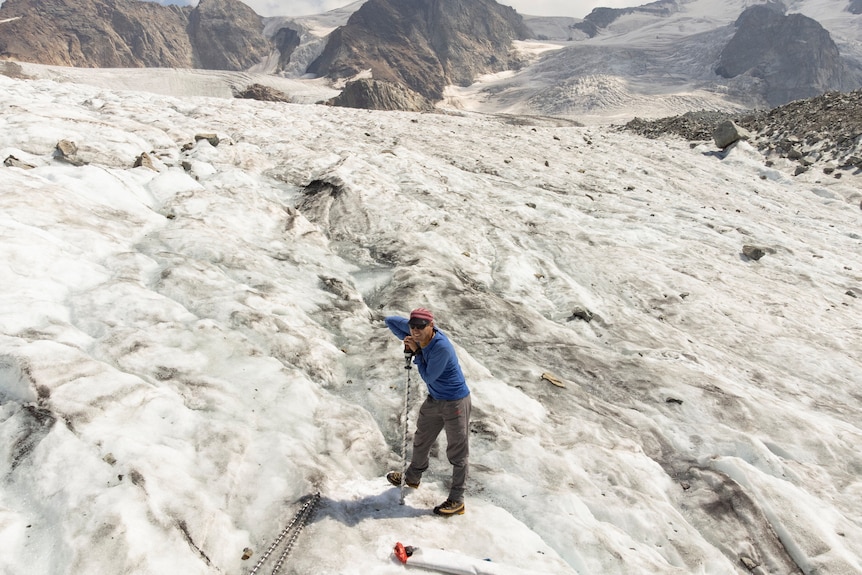 A man is standing on a mountain covered in snow while leaning on a walking pole. 