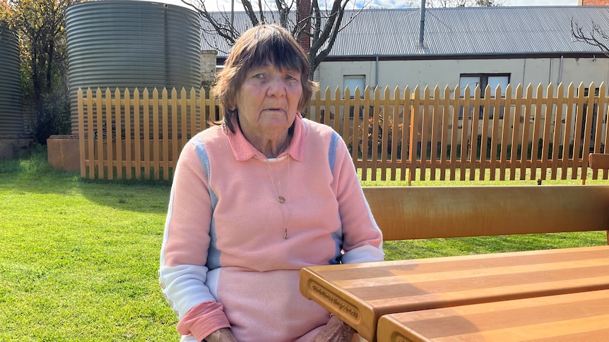 An older Indigenous woman sits at a table in a grassy yard.