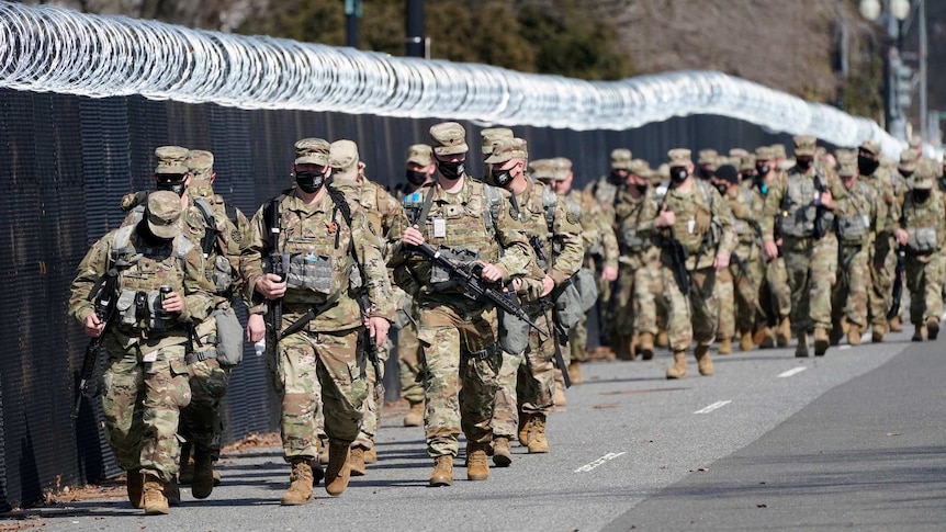 US National Guard in uniform and black masks holding rifles walk along a large black fence with barbed wire on top