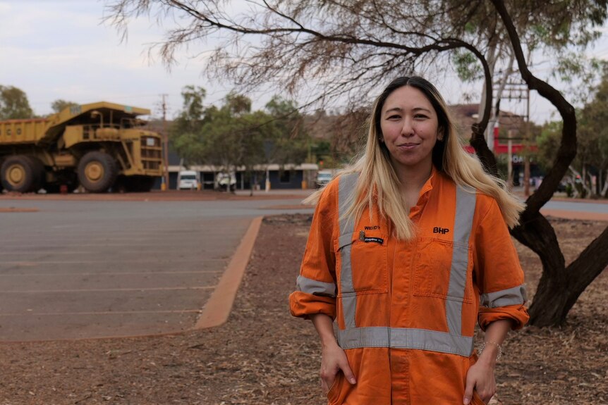 A woman wearing high-vis clothing stands in front of trees and a carpark with a large mining truck in the background.