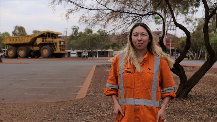A woman wearing high-vis clothing stands in front of trees and a carpark with a large mining truck in the background.