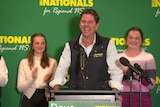 A smiling man stands at a podium flanked by two young girls applauding.