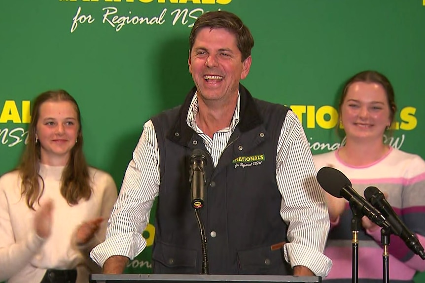 A smiling man stands at a podium flanked by two young girls applauding.