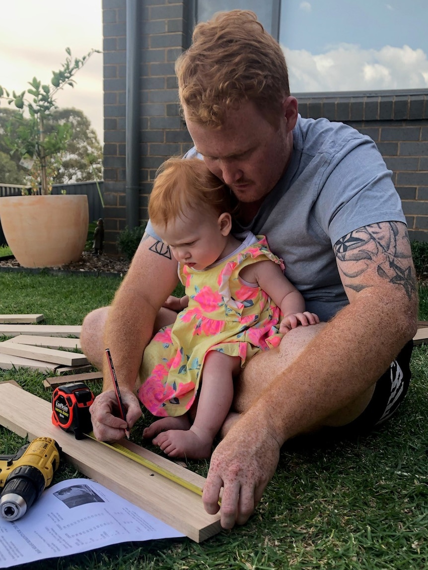 A man sits on the ground with a young girl in his lap, measuring out slabs of wood around her.