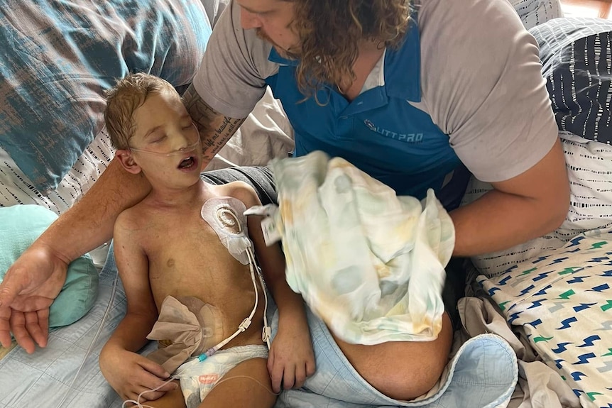 A small boy attached to medical monitoring equipment sits on a bed