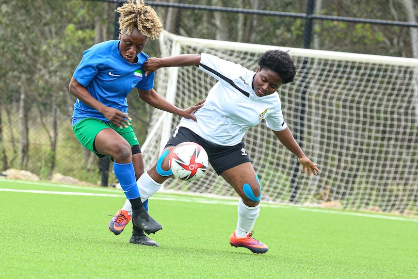 Two women are playing football, they have their arms on each other as they both attempt to get to the ball.