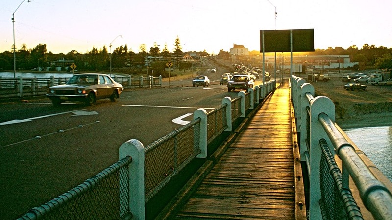 At dusk, you view golden sunlight bouncing off of wooden slats on a pedestrian and road bridge over a river.