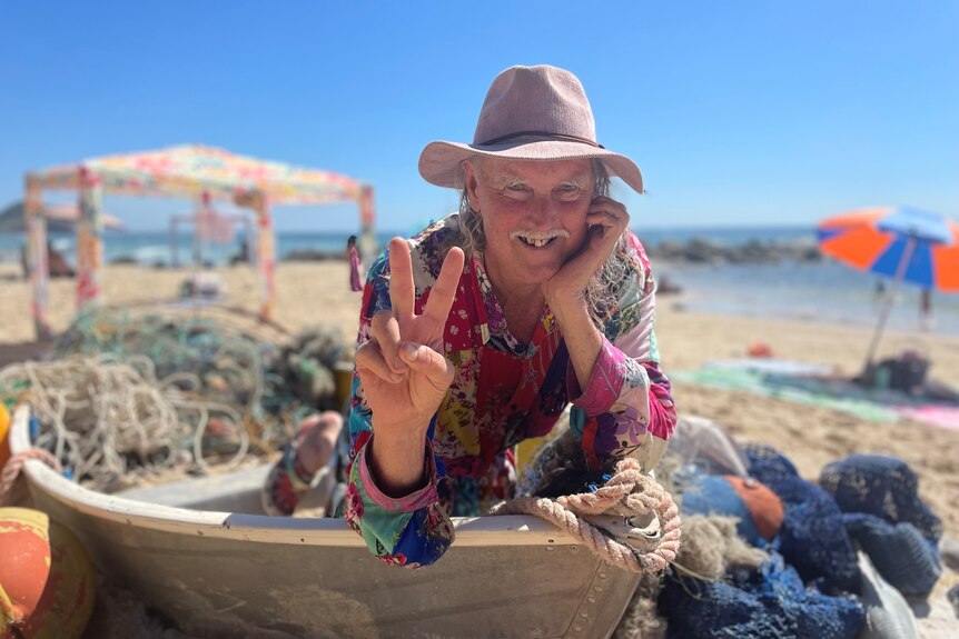 a man in a rainbow top gives the peace sign while sitting inside a dinghy sculpture on the beach