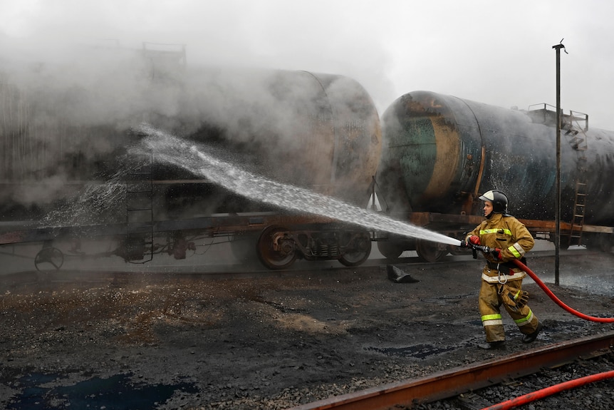 A firefighter points a water hose at a burnt tank car on train tracks, amid smoke and steam.