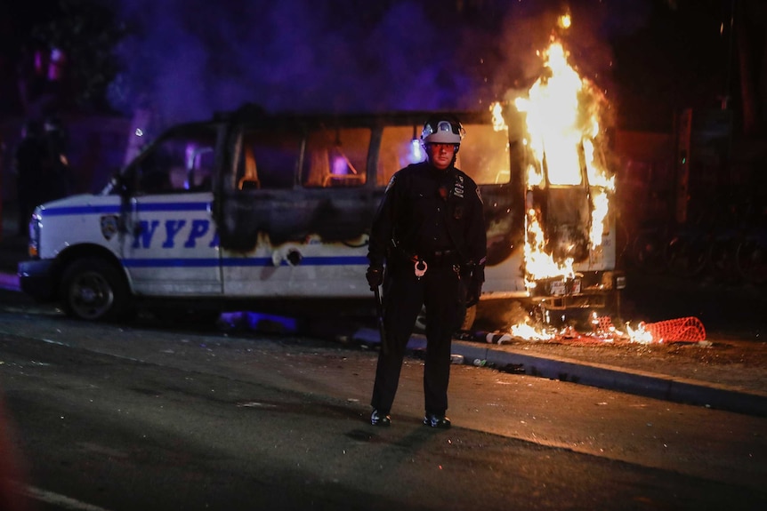 A police officer in a helmet stands in front of a burning NYPD van.