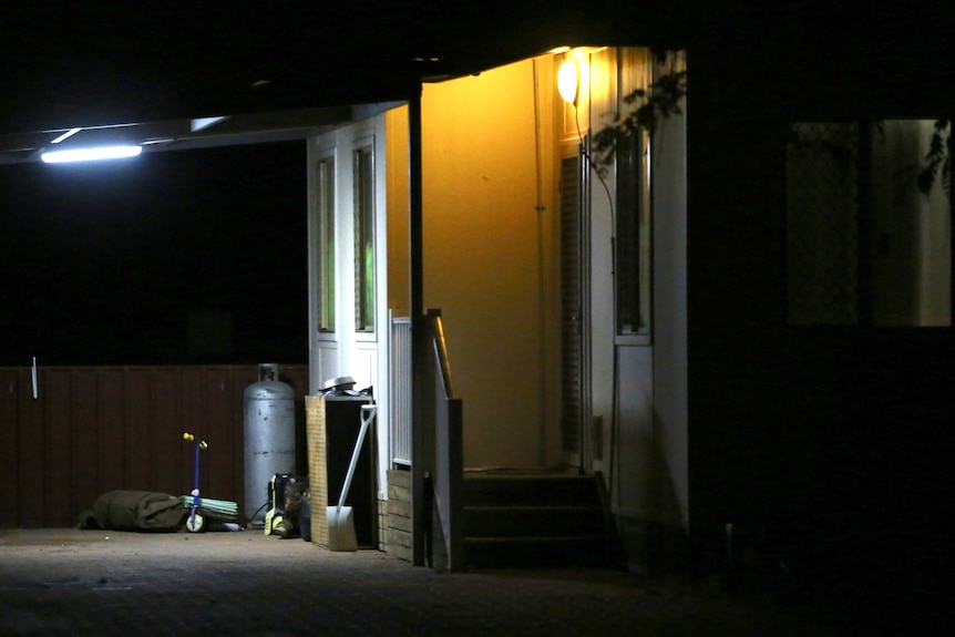 A close-up shot of the side of a house at night with items including a gas bottle, shovel and scooter outside.