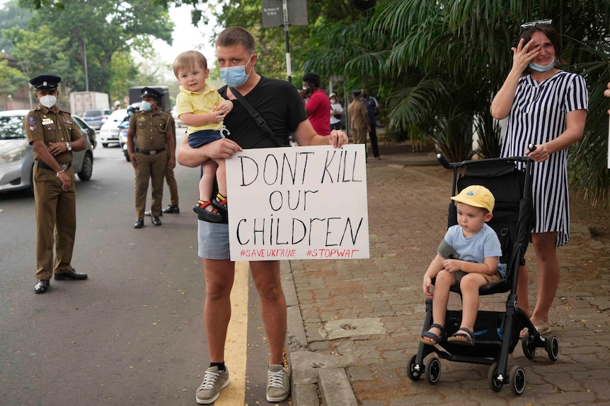 A man holds a child and a sign which reads 'Don't kill our children', next to a crying woman with a child in a pram