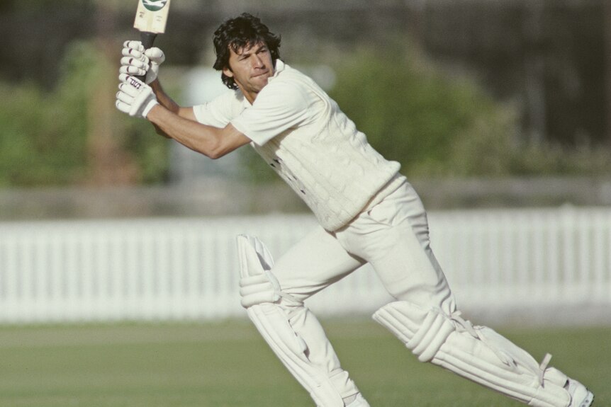 Imran Khan leans over as he swings his bat on a cricket pitch.