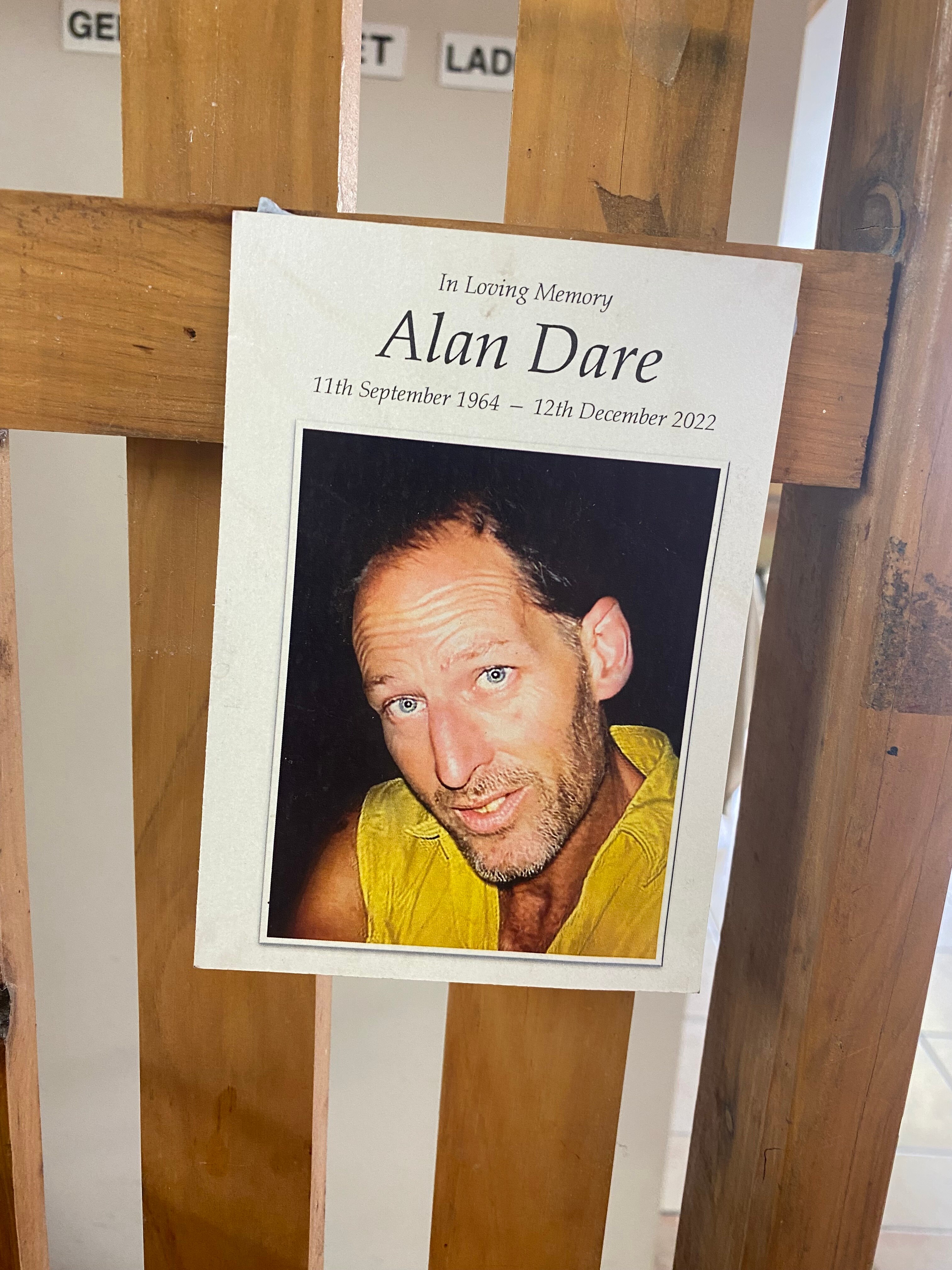 a poster hanging on a barrier that says 'in loving memory alan dare' with a photo of him wearing a yellow shirt