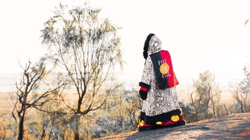 Picture of a man in the bush wearing a long coat with the Aboriginal flag on the back of it.