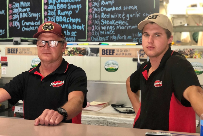 Two men in black shirts with caps on their heads, standing in a butcher shop