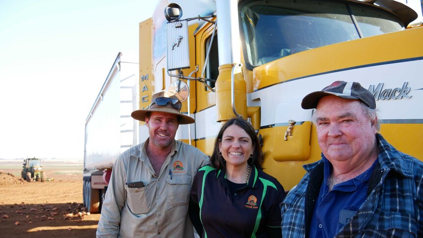 Two men and a woman stand in front of a yellow Mack truck.