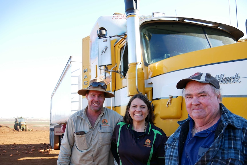 Two men and a woman stand in front of a yellow Mack truck.