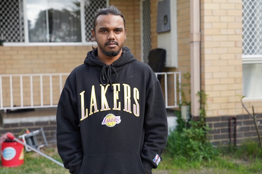 A man wearing a black hoodie stands outside a house.