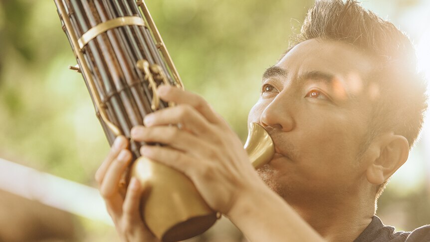 A close-up photo of Wo Tong playing the sheng. Blurred trees are in the background.