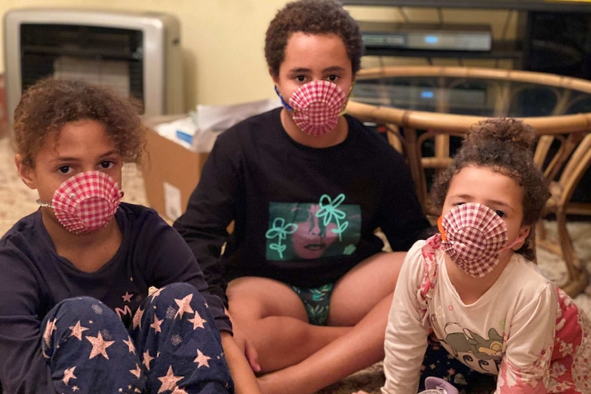 Three children sit in a lounge room wearing face mask they have fashioned out of paper muffin pans.