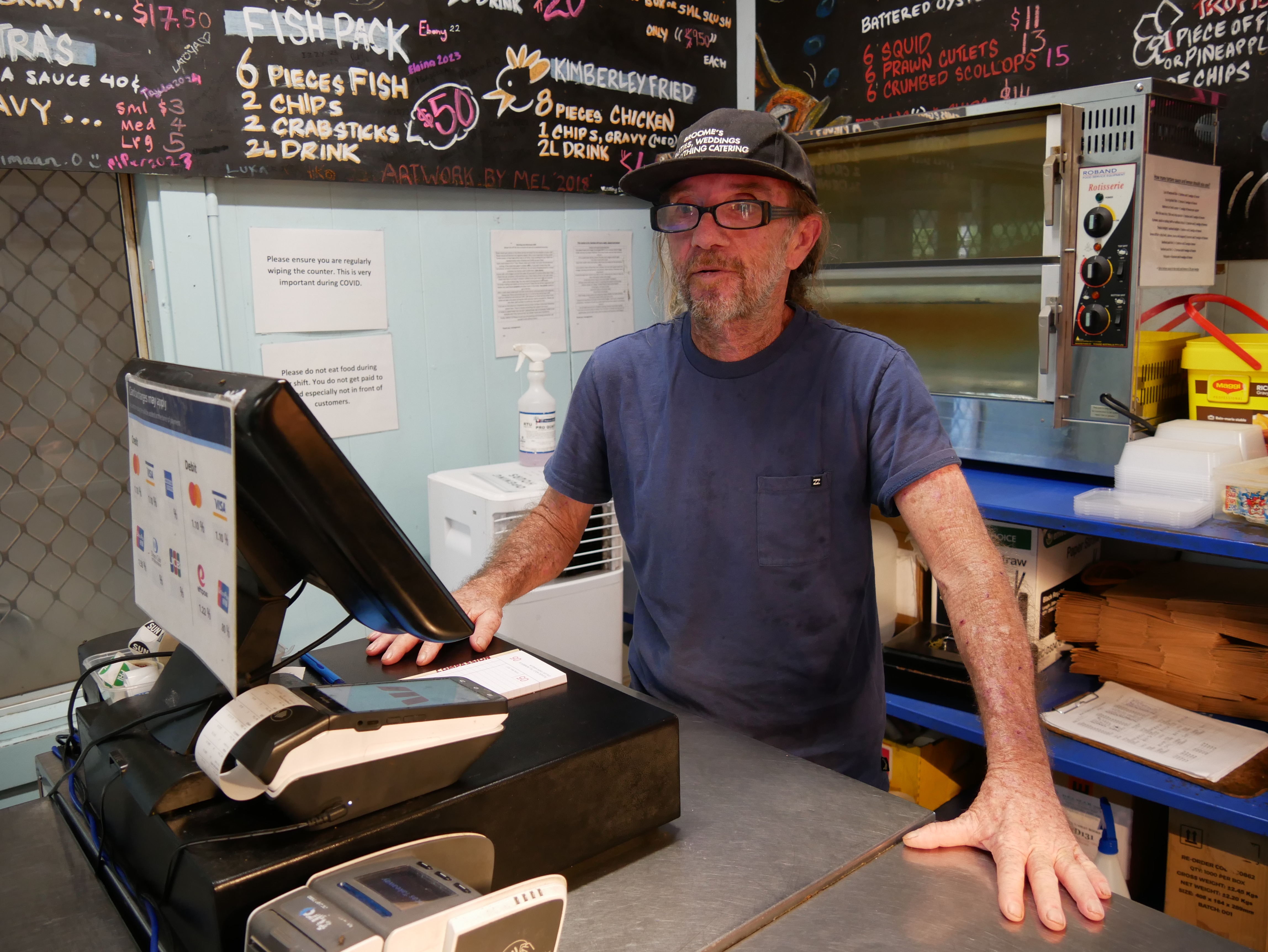 Bearded man wearing black hat and glasses, standing behind money till in food store