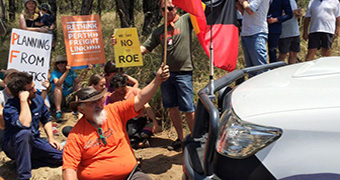 Protestors carrying placards sit in front of a vehicle in bushland.